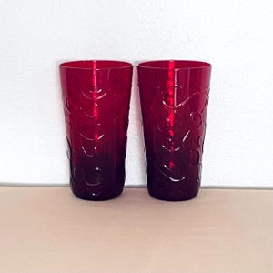 May include: Two tall, ruby red drinking glasses with a decorative, swirling pattern. The glasses are made of a translucent material, and the color darkens towards the base. The glasses are displayed against a white wall and a light brown surface.