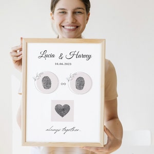 Puede incluir: Una impresión enmarcada con fondo blanco y texto negro. El texto dice "Lucia & Harvey 10.06.2023" y "siempre juntos". La impresión presenta dos diseños de huellas dactilares en círculos y un diseño de huella dactilar en forma de corazón.