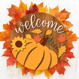 Op de afbeelding: Een houten welkomstbord met een pompoen- en zonnebloemontwerp. Het bord is omgeven door herfstbladeren in tinten oranje, rood en geel. Het woord "welcome" staat in een sierlijk lettertype bovenaan het bord.
