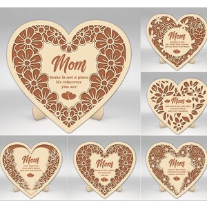 Op de afbeelding: Een verzameling houten hartvormige plaquettes met bloemen- en tekstontwerpen. Elke plaquette bevat het woord "Mom" en een unieke boodschap. De plaquettes worden weergegeven op kleine houten standaards. De tekst bevat zinnen als "thuis is geen plaats".