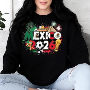 K&ouml;nnte beinhalten: Schwarzes Sweatshirt mit einem farbenfrohen Grafikdesign. Das Design enth&auml;lt das Wort "MEXICO" in Gr&uuml;n und Wei&szlig;, die Zahl "2026", einen Fu&szlig;ball, einen Sombrero und eine Troph&auml;e. Feuerwerk und Farbspritzer vervollst&auml;ndigen das Design.