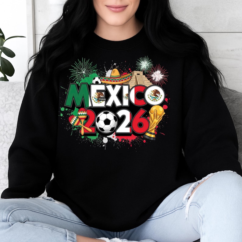 K&ouml;nnte beinhalten: Schwarzes Sweatshirt mit einem farbenfrohen Grafikdesign. Das Design enth&auml;lt das Wort "MEXICO" in Gr&uuml;n und Wei&szlig;, die Zahl "2026", einen Fu&szlig;ball, einen Sombrero und eine Troph&auml;e. Feuerwerk und Farbspritzer vervollst&auml;ndigen das Design.