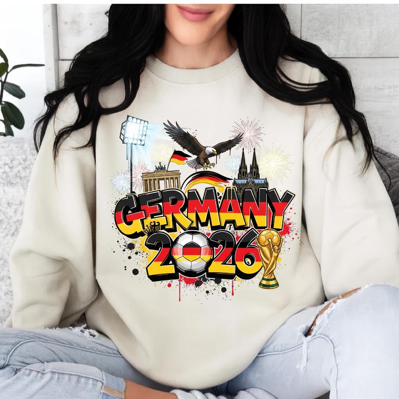 K&ouml;nnte beinhalten: Cremefarbenes Sweatshirt mit farbenfrohem Grafikdesign "GERMANY 2026". Das Design beinhaltet einen Fu&szlig;ball, eine Troph&auml;e, einen Adler und deutsche Wahrzeichen. Das Design ist mit Feuerwerk akzentuiert.