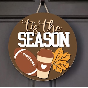 May include: Round wooden door sign with the text "'tis the SEASON" in white and tan. The sign features a football, coffee cup with a heart, and a maple leaf in fall colors. A jute rope is attached for hanging.