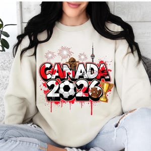 K&ouml;nnte beinhalten: Cremefarbenes Sweatshirt mit einem grafischen Design mit dem Text "CANADA 2026" in Rot, Wei&szlig; und Schwarz. Das Design umfasst einen Fu&szlig;ball, eine Troph&auml;e, einen Biber und Feuerwerk.