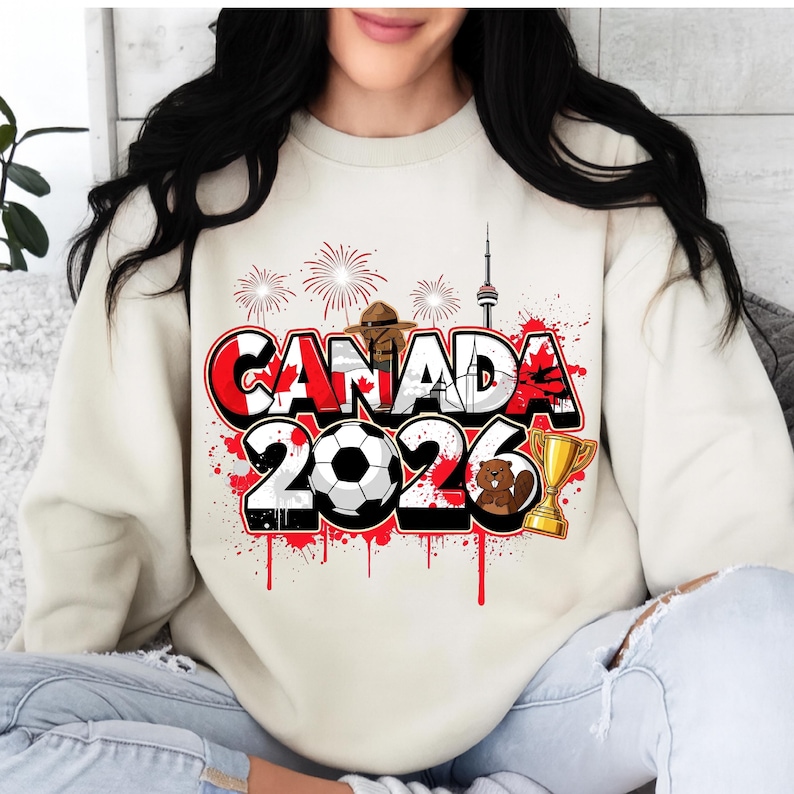 K&ouml;nnte beinhalten: Cremefarbenes Sweatshirt mit einem grafischen Design mit dem Text "CANADA 2026" in Rot, Wei&szlig; und Schwarz. Das Design umfasst einen Fu&szlig;ball, eine Troph&auml;e, einen Biber und Feuerwerk.