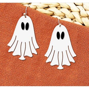 May include: A pair of white ghost earrings with black oval eyes and a flowing bottom design. The earrings are displayed on an orange fabric background, suggesting a Halloween theme. The earrings are hanging from silver hooks.