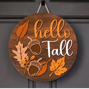 Könnte beinhalten: Runder Holzschild mit den Worten "hello Fall" in orange und weißer Schrift. Das Schild ist mit orangefarbenen und braunen Blättern und Eicheln verziert und wird an einem Seil aufgehängt.