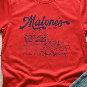 Op de afbeelding: Rood t-shirt met een marineblauwe afbeelding van een hamburger en friet, met de tekst "Malone's since 2015" en "Proudly Serving Briar University". Het shirt heeft korte mouwen.