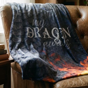 May include: A plush, dark blue blanket draped over a brown leather armchair. The blanket features the text "my DRAGON awaits" in a decorative font. The bottom of the blanket has a fiery orange and yellow design.