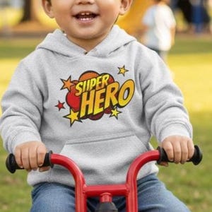 May include: A young child wearing a gray hoodie with the words "SUPER HERO" in a comic book style graphic, riding a red tricycle on a grassy lawn. The child is smiling. The background shows a playground and trees.