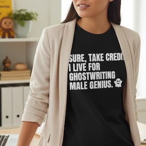 May include: Black t-shirt with white text that reads "Sure, take credit, I live for ghostwriting male genius." A small ghost graphic is next to the text. The person is wearing a light tan blazer and black pants.
