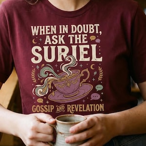 Può includere: T-shirt bordeaux con la scritta "WHEN IN DOUBT, ASK THE SURIEL" e una grafica di una tazza da tè con vapore. La maglietta ha anche le parole "GOSSIP AND REVELATION". Una persona tiene in mano una tazza.