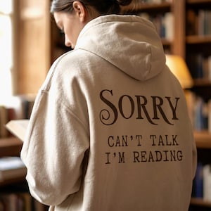May include: A beige hooded sweatshirt featuring dark brown text printed on the back. The text reads, "SORRY CAN'T TALK I'M READING" in a combination of elegant serif and block lettering. The garment includes a hood and long sleeves.