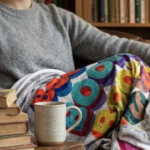 May include: A gray sweater and a colorful blanket with a geometric design. A stack of old books and a speckled mug with coffee are visible. The blanket features a pattern of large, colorful shapes.
