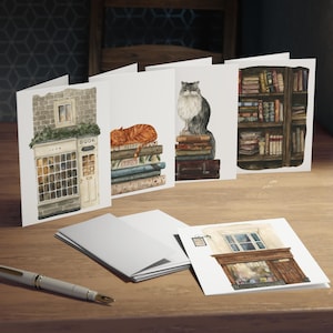 May include: A collection of watercolor greeting cards featuring literary themes. The cards depict a bookstore, a cat sleeping on books, a fluffy cat atop books, and a bookshelf. The cards are arranged on a wooden surface with a pen.