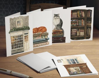 May include: A collection of watercolor greeting cards featuring literary themes. The cards depict a bookstore, a cat sleeping on books, a fluffy cat atop books, and a bookshelf. The cards are arranged on a wooden surface with a pen.