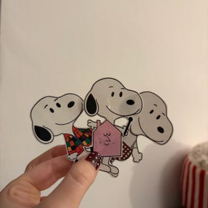 May include: A set of three Snoopy stickers. The Snoopy stickers are white with black ears and a black nose. One Snoopy is wearing a plaid outfit. Another Snoopy is holding a pink house with the number 31 on it.