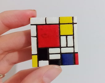 May include: A small, rectangular wooden block with a colourful abstract design. The design includes a red square, yellow and blue rectangles, and black and white accents, similar to a Mondrian painting. The block is held by a hand.