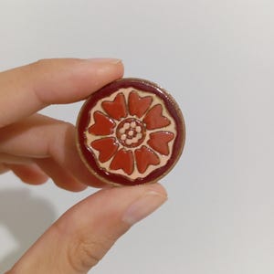 May include: A round, red and cream-colored pin with a floral design. The pin features a central flower with heart-shaped petals and a dark red border. The pin is held between fingers.