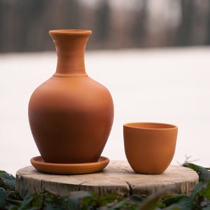 May include: A terracotta-colored ceramic jug with a rounded body and a long, slender neck, accompanied by a matching cup. The jug sits on a small, round saucer. The items are displayed on a wooden surface against a blurred outdoor backdrop.