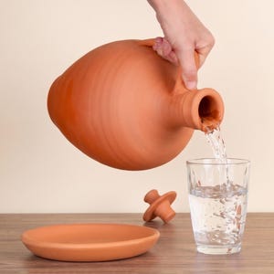 May include: A terracotta pitcher with a rounded body and a handle is tilted to pour clear water into a glass. A matching terracotta lid and a circular terracotta saucer are positioned nearby on a flat surface, completing the set.