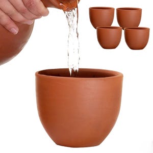 May include: A set of five terracotta-colored clay cups with a smooth, rounded shape. One cup is positioned in the foreground while water is being poured into it from a matching pitcher, and four additional identical cups are arranged in the background.
