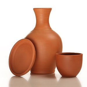 Puede incluir: Un juego de cerámica de terracota que incluye una jarra alta y redondeada con cuello estrecho, una tapa a juego y una taza pequeña y redondeada. Los artículos son de un color marrón anaranjado cálido y terroso, sobre fondo blanco.