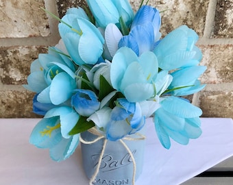 May include: A bouquet of light blue and white artificial tulips in a gray Mason jar. The jar has the word "Ball MASON" printed on it and is tied with twine. The flowers are arranged against a brick wall.