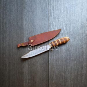 Può includere: Un coltello fatto a mano con una lama a motivi e un manico in legno a strisce marroni e beige. È accompagnato da un fodero in pelle marrone. La parola "Sitecture" è stampata sull'immagine.
