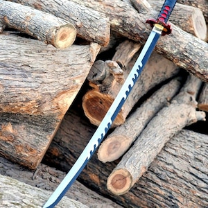 May include: A katana sword with a black and red handle, a gold handguard, and a silver blade with a wavy design along the edge. The sword is resting on a pile of wooden logs.