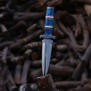 May include: A handcrafted knife with a silver blade and a blue, white, and red handle. The knife is set against a backdrop of stacked wood logs. The handle features a layered design with a decorative bolster.