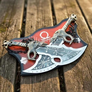 Puede incluir: Un adorno de pared decorativo con dos hachas plateadas con diseños intrincados y mangos con forma de dragón. Las hachas están montadas sobre una placa roja y negra en forma de escudo con un emblema circular. El texto "Sitecture" es visible.