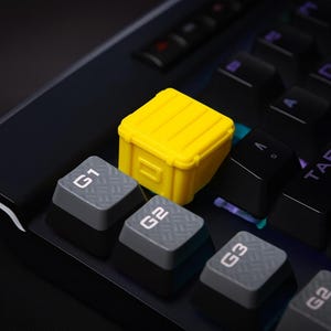 May include: A close-up of a mechanical keyboard with a yellow, miniature crate keycap. The keyboard has black keys with white lettering and gray keys labeled G1, G2, and G3. The keyboard is on a black surface.
