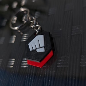 May include: A black and red keychain with a white fist symbol. The keychain is shaped like a hexagon.