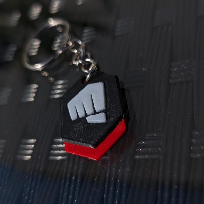 May include: A black and red keychain with a white fist symbol. The keychain is shaped like a hexagon.