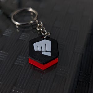 May include: A black and red keychain with a white fist symbol. The keychain is in the shape of a hexagon.