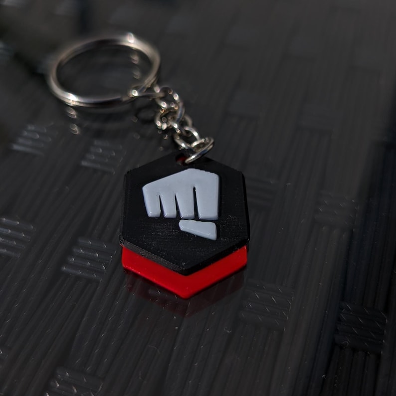 May include: A black and red keychain with a white fist symbol. The keychain is in the shape of a hexagon.