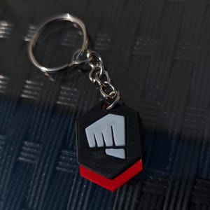 May include: A black and red keychain with a white fist logo. The keychain is in the shape of a hexagon.