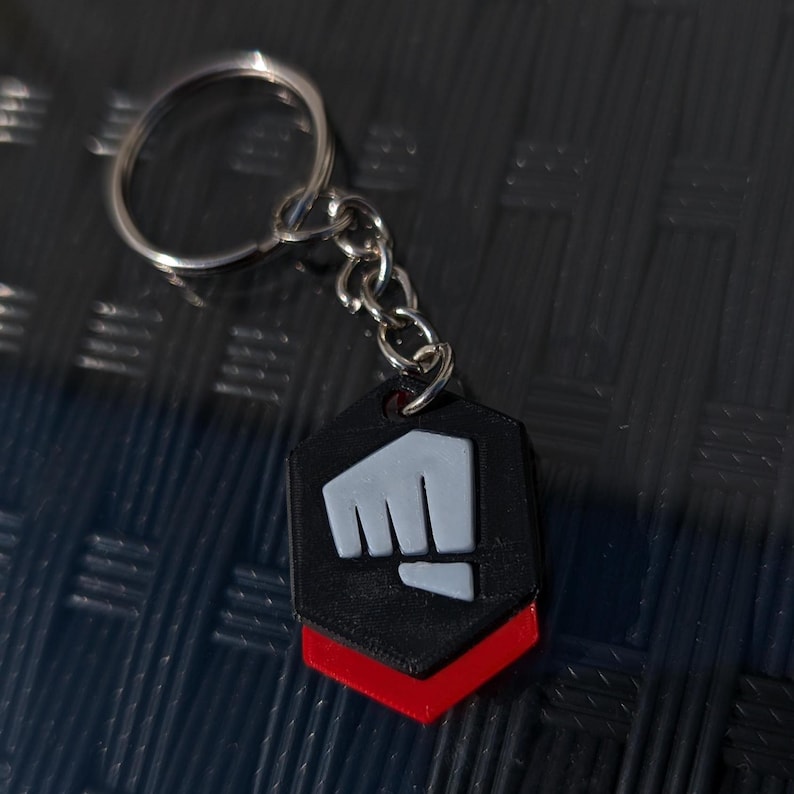 May include: A black and red keychain with a white fist logo. The keychain is in the shape of a hexagon.