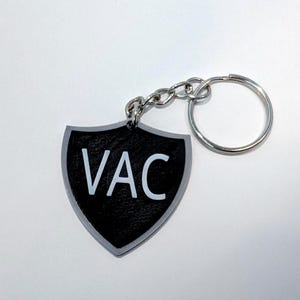 Puede incluir: Un llavero negro en forma de escudo con las letras "VAC" en blanco. El llavero tiene un borde plateado, una cadena plateada y un anillo plateado. El fondo es blanco.