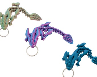 Pode incluir: Três chaveiros em forma de dragão em diferentes cores: azul-esverdeado, roxo e verde. Cada chaveiro tem um anel prateado.