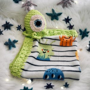 Peut inclure: Un jouet monstre en crochet vert avec un gros œil attaché à une couverture rayée blanche et verte avec un imprimé monstre.