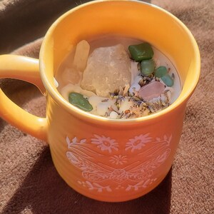 Puede incluir: Una taza de cerámica amarilla con un diseño floral blanco, llena de una vela blanca y varios cristales, incluyendo cuarzo transparente, aventurina verde y cuarzo rosa.