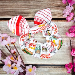 May include: White and red fabric hair scrunchie with a repeating pattern of In-N-Out Burger food items. The scrunchie is tied in a bow.