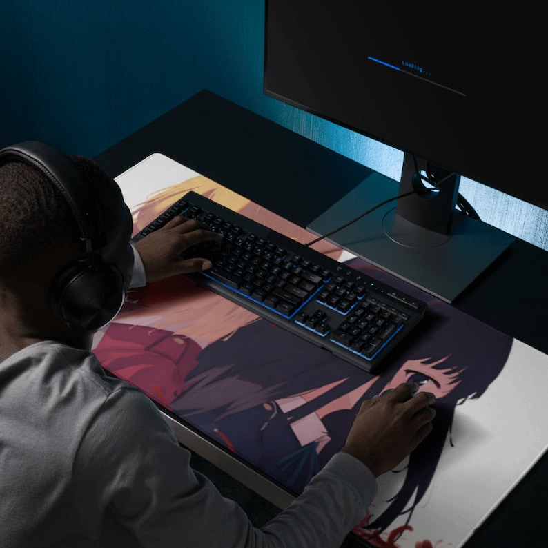May include: A person using a black keyboard with blue backlit keys and a mouse pad featuring an anime illustration of two women. The computer monitor in the background is displaying a loading screen.