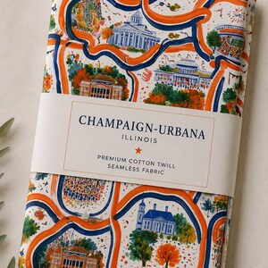 May include: A folded piece of white cotton twill fabric featuring a colorful illustrated map pattern of Champaign-Urbana, Illinois, with orange and blue winding lines, trees, and buildings. A paper band reads: CHAMPAIGN-URBANA ILLINOIS PREMIUM COTTON TWILL SEAMLESS FABRIC.