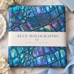Könnte beinhalten: Ein gefaltetes Stoffstück mit einem blauen, holografischen Krokodilledermuster mit schimmernden violetten und türkisfarbenen Akzenten. Ein weißes Papierband umschließt die Mitte mit dem aufgedruckten Text BLUE HOLOGRAPHIC und COMBED COTTON PRIMA.