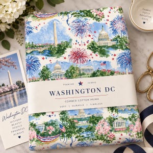 Könnte beinhalten: Ein gefaltetes Stück weißer Stoff mit einem Aquarelldruck von Wahrzeichen aus Washington DC, darunter das Kapitol und das Washington Monument, mit roten und blauen Feuerwerken und Sternen. Eine Papierbanderole trägt die Aufschrift WASHINGTON DC.