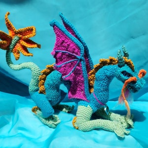 May include: A crocheted dragon toy with a turquoise body, pink wings, and orange and yellow accents. The dragon has a long tail and a friendly expression.